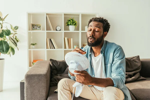 Homme afro-américain tenant souffler ventilateur électrique tout en souffrant de chaleur à la maison — Photo de stock