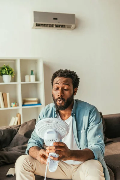 Handsome african american man holding blowing electric fan while sitting on sofa and suffering from heat at home — Stock Photo