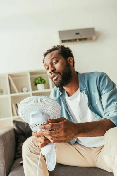 Épuisé homme afro-américain tenant souffler ventilateur électrique tout en étant assis sur le canapé et souffrant de chaleur à la maison — Photo de stock