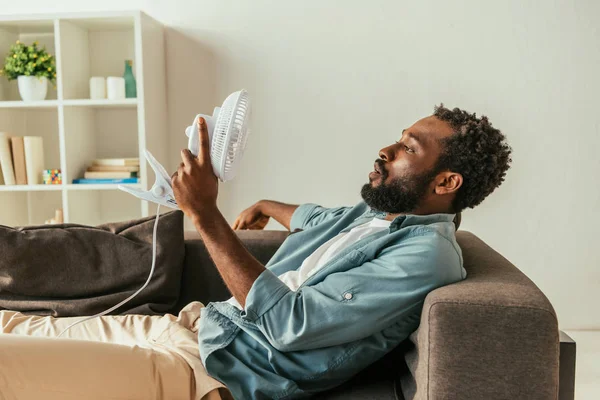 Cansado afro-americano homem deitado no sofá com ventilador elétrico soprando enquanto sofre de calor de verão — Fotografia de Stock