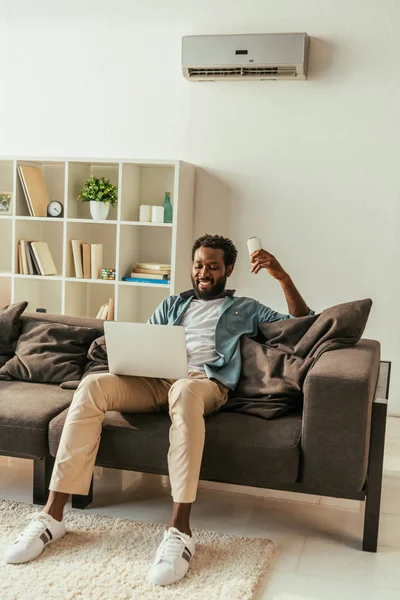 Allegro uomo afroamericano utilizzando il computer portatile e tenendo il telecomando del condizionatore d'aria mentre si siede sul divano a casa — Foto stock