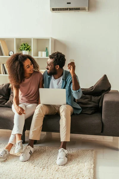 Cheerful african american man using laptop and holding air conditioner remote controller while sitting on sofa near smiling african american woman — Stock Photo