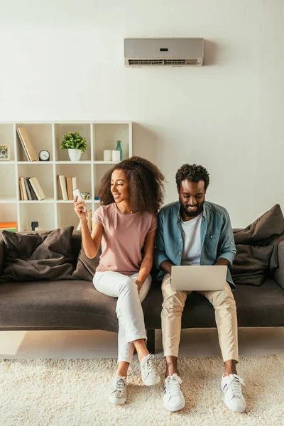 Smiling african woman holding air conditioner remote controller while sitting on couch near african american man using laptop — Stock Photo