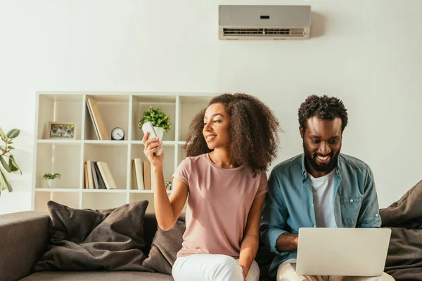 Smiling african american man using laptop while sitting on sofa near cheerful african american woman holding air conditioner remote controller — Stock Photo