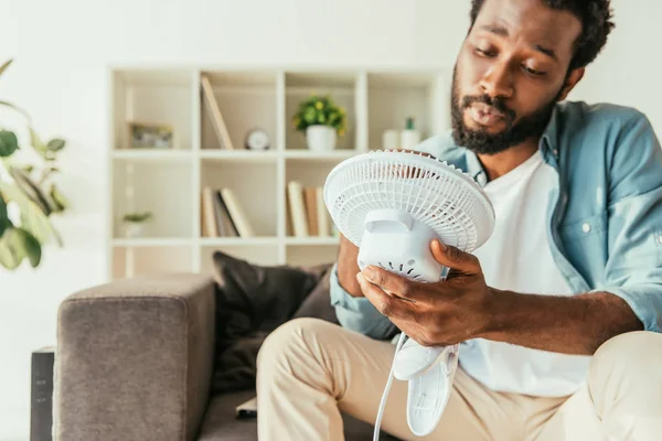Exhausted african american man holding blowing electric fan while suffering from heat at home — Stock Photo
