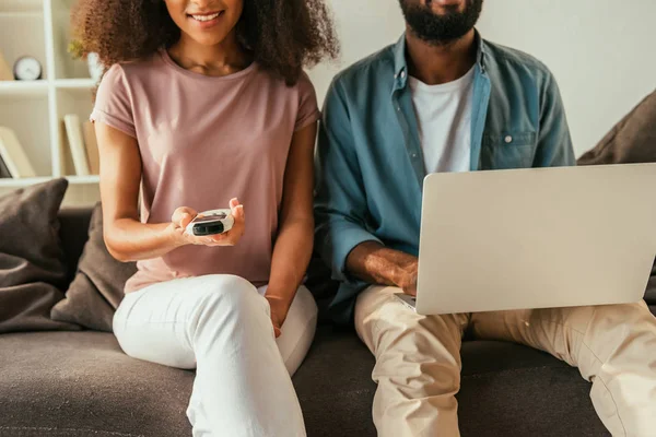 Vue partielle de l'homme afro-américain en utilisant un ordinateur portable tout en étant assis sur le canapé près de la femme afro-américaine tenant climatiseur télécommande — Photo de stock