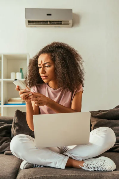 Dissatisfied african american woman holding air conditioner remote controller while sitting on sofa with laptop — Stock Photo