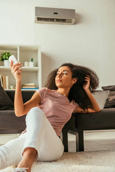 Dissatisfied african american woman sitting on floor at home and using air conditioner remote controller — Stock Photo