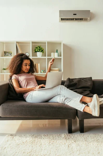 Mujer americana africana bonita usando el ordenador portátil y sosteniendo el regulador remoto del aire acondicionado mientras que miente en sofá en el país - foto de stock