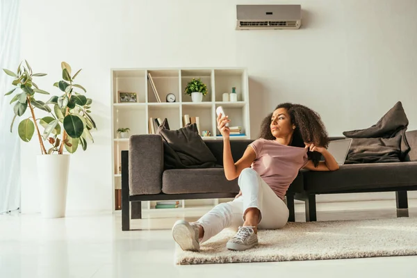 Atractiva mujer afroamericana sentada en el suelo y usando el controlador remoto de aire acondicionado mientras sufre de calor de verano en casa - foto de stock