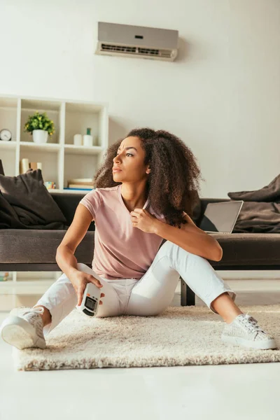 Mujer afroamericana joven mirando hacia otro lado y sosteniendo el controlador remoto del aire acondicionado mientras está sentada en el sofá en casa - foto de stock