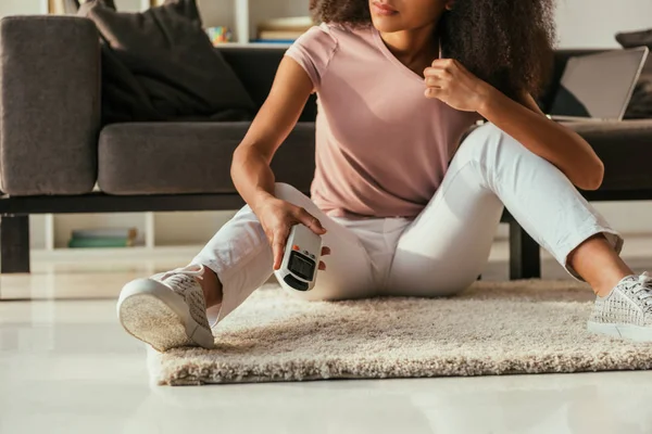 Vista ritagliata della donna afroamericana seduta sul pavimento e con in mano il telecomando del condizionatore d'aria — Foto stock