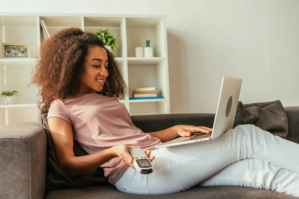 Усміхнена афроамериканська жінка використовує ноутбук і тримає пульт дистанційного керування кондиціонера, лежачи на дивані і використовуючи ноутбук — стокове фото