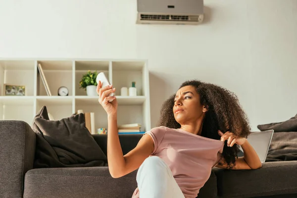 Dissatisfied african american woman sitting on sofa and using air conditioner remote controller while suffering from summer heat at home — Stock Photo