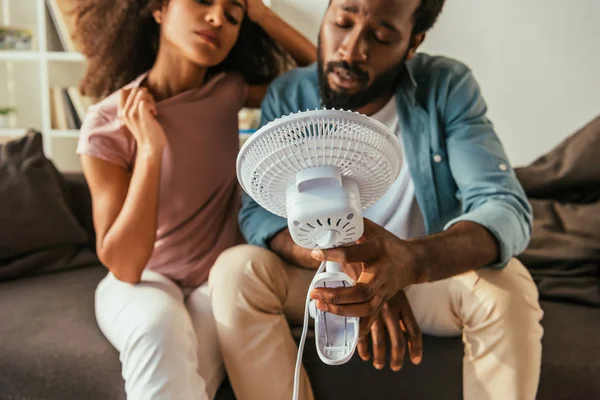 Épuisé afro-américain homme et femme tenant souffler ventilateur électrique tout en souffrant de chaleur estivale — Photo de stock