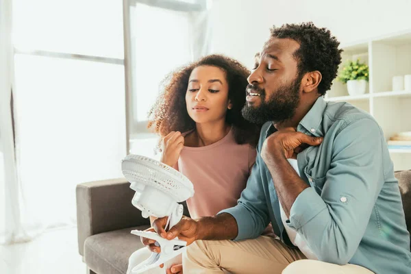 Joven africano hombre y mujer que sufre de calor de verano y la celebración de soplado ventilador eléctrico - foto de stock