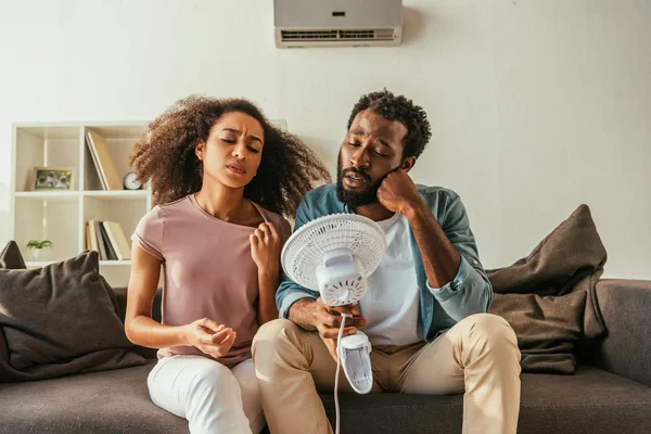 Infeliz afroamericano hombre y mujer sosteniendo soplado acondicionador eléctrico mientras que sufre de calor de verano en casa - foto de stock