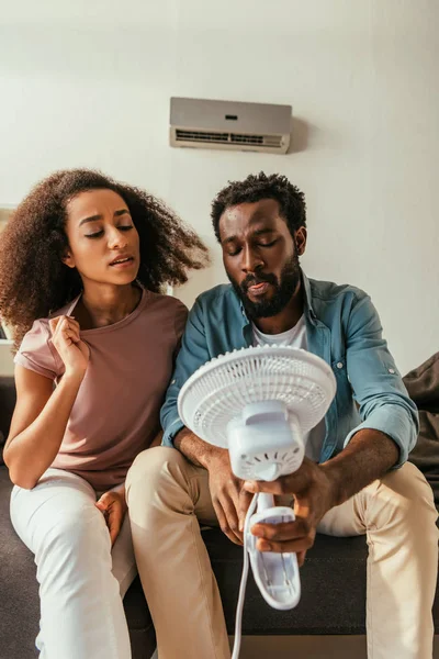 Hombre y mujer afroamericanos insatisfechos sentados en el sofá y sosteniendo ventilador eléctrico que sopla - foto de stock
