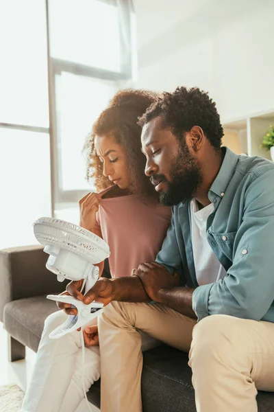 Afrikanisch-amerikanischer Mann und Frau leidet unter Sommerhitze zu Hause und hält blasenden elektrischen Ventilator — Stockfoto
