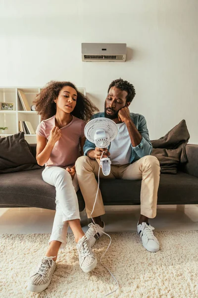 Hombre y mujer afroamericanos jóvenes sentados en el sofá y sosteniendo ventilador eléctrico mientras sufren de calor en casa - foto de stock