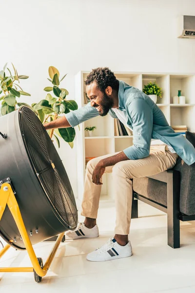 Homme afro-américain joyeux assis devant ventilateur électrique tout en souffrant de chaleur à la maison — Photo de stock