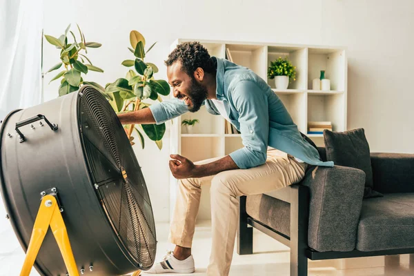 Excited african american man sitting near electric ventilator at home — Stock Photo