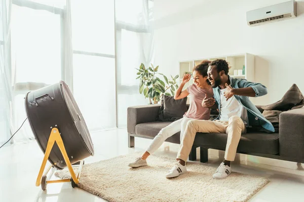 Excited african american man and woman sitting on sofa in front of blowing electric fan — Stock Photo
