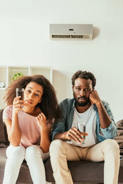 Infeliz afroamericano hombre y mujer sentado en el sofá y beber agua mientras que sufre de calor de verano en casa - foto de stock