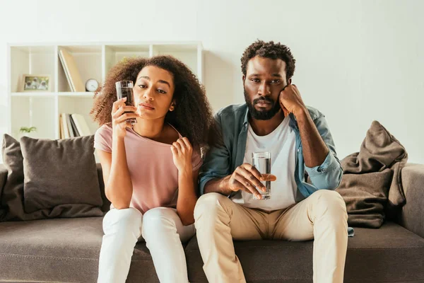 Esgotado afro-americano homem e mulher bebendo água de óculos enquanto sentado no sofá e sofrendo de calor de verão em casa — Fotografia de Stock