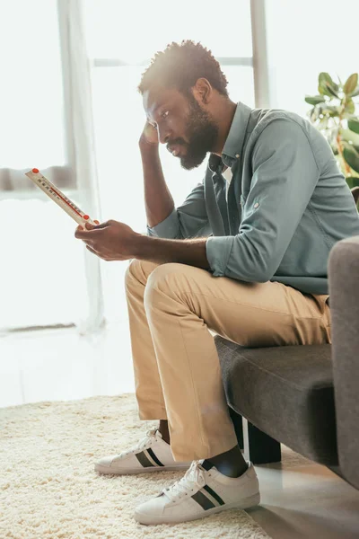 Unhappy african american man sitting on sofa and holding thermometer while suffering from summer heat at home — Stock Photo