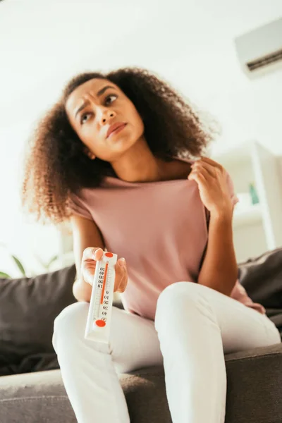 Exhausted african american woman suffering from summer heat while sitting on sofa and holding thermometer showing high temperature — Stock Photo