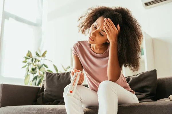 Mujer afroamericana infeliz mirando el termómetro que muestra alta temperatura y sosteniendo la mano en la cabeza - foto de stock