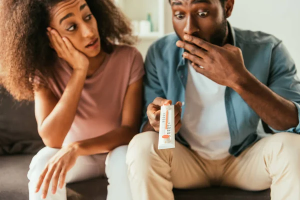 Sorprendido africano americano hombre celebración termómetro mostrando alta temperatura mientras sentado cerca sorprendido africano americano mujer - foto de stock