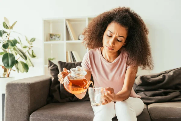 Jeune femme afro-américaine verser le thé dans une tasse tout en étant assis sur le canapé et souffrant de chaleur à la maison — Photo de stock