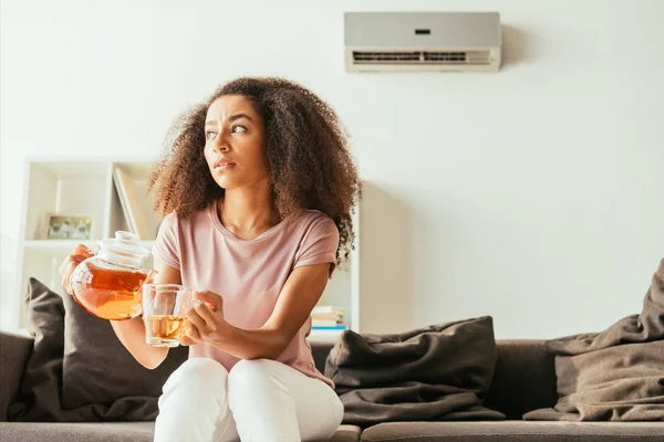 Mulher afro-americana infeliz derramando chá na xícara e olhando para longe enquanto sentado no sofá e sofrendo de calor de verão — Fotografia de Stock