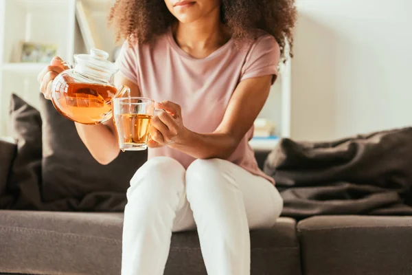 Teilansicht einer afrikanisch-amerikanischen Frau, die auf dem Sofa sitzt und Tee in Tasse gießt — Stockfoto