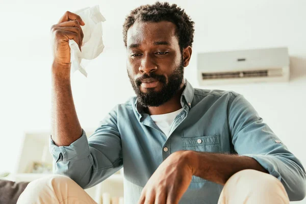 Unhappy african american man holding napkin while suffering from summer heat at home — Stock Photo