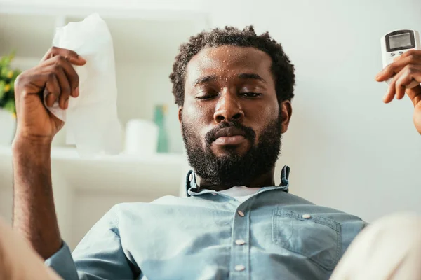 Homem americano africano com rosto suado segurando guardanapo e controle remoto enquanto sofre de calor em casa — Fotografia de Stock