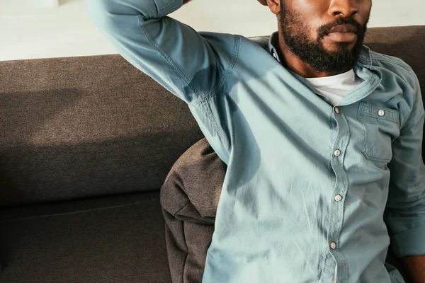 Vista parcial del hombre afroamericano en camisa sudorosa sentado en el sofá y sufriendo de calor - foto de stock