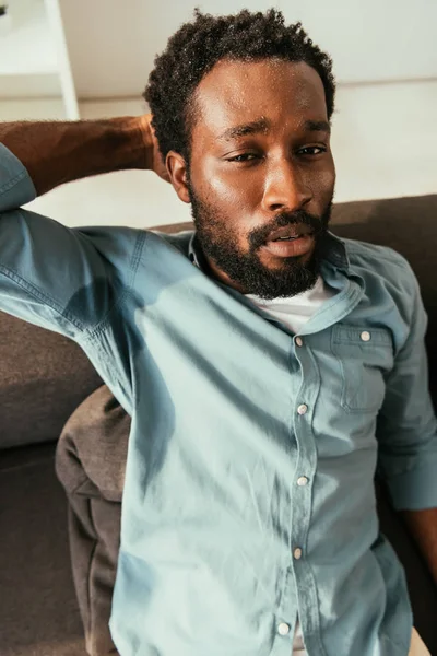 Unhappy african american man in sweaty shirt sitting on couch and suffering from summer heat — Stock Photo