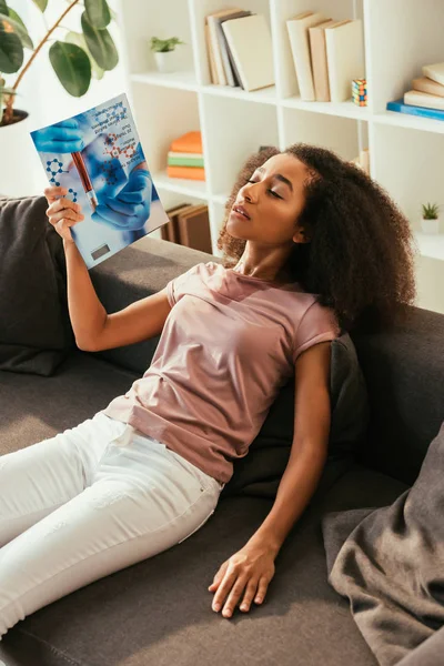 Exhausted african american woman waving with magazine while sitting on sofa with closed eyes and suffering from summer heat — Stock Photo