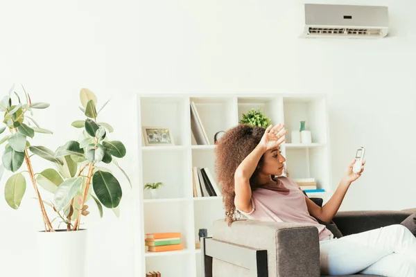 Young african american woman using remote controller while sitting on sofa under air conditioner — Stock Photo