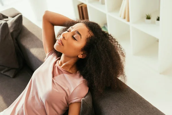 Mujer afroamericana agotada sentada en el sofá con los ojos cerrados y sufriendo de calor - foto de stock