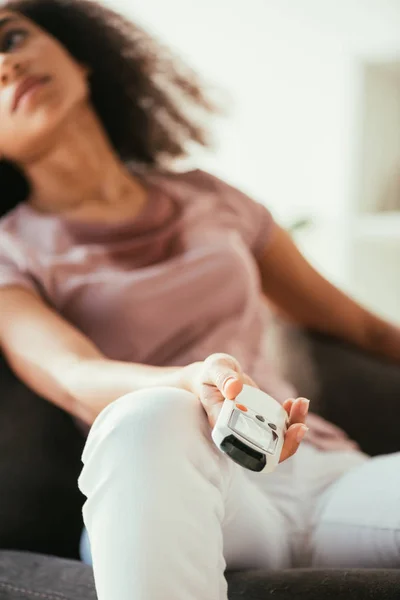 Foco seletivo da mulher afro-americana esgotada segurando controlador remoto de ar condicionado enquanto sofre de calor de verão em casa — Fotografia de Stock