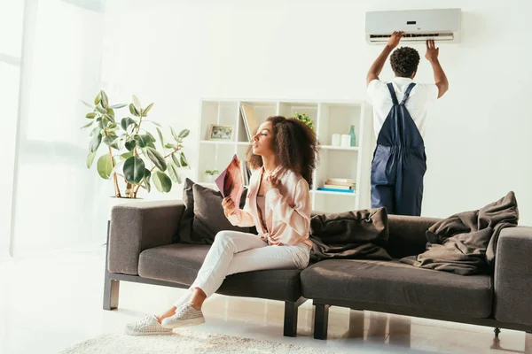 Mujer afroamericana bonita sentada en el sofá y saludando con la revista, mientras que el reparador afroamericano fija el aire acondicionado - foto de stock