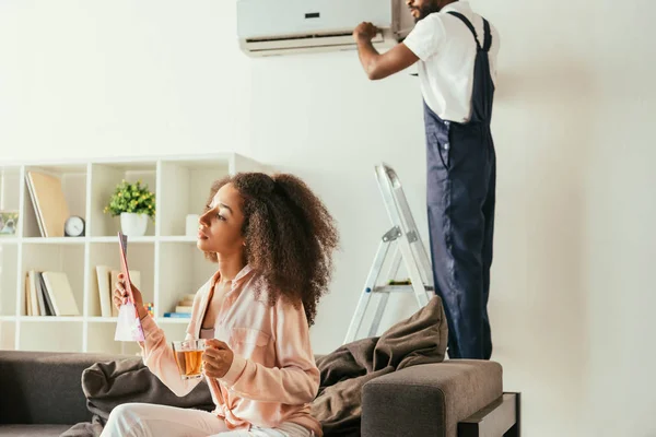 Femme afro-américaine réfléchie assis sur le canapé avec magazine et tasse de thé, tandis que l'homme à tout faire afro-américain réparer climatiseur — Photo de stock