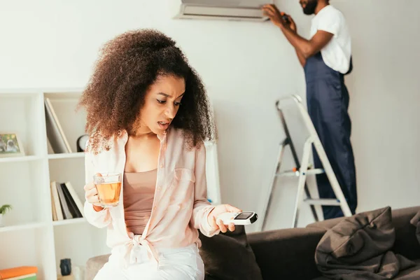 Dissatisfied african american woman looking at remote controller, while african american repairman fixing air conditioner — Stock Photo