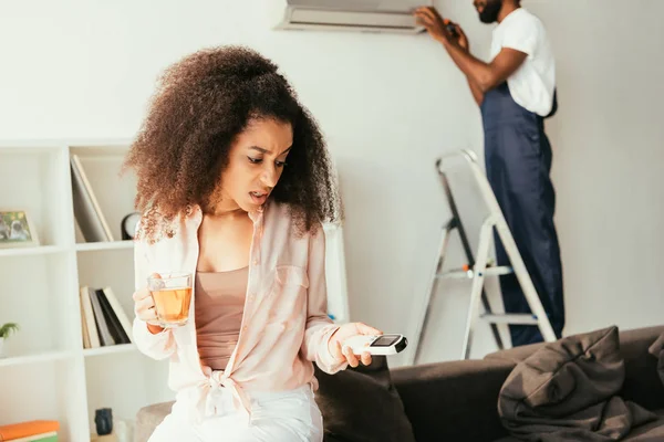 Mulher afro-americana insatisfeita olhando para controlador remoto, enquanto reparador afro-americano fixando ar condicionado — Fotografia de Stock