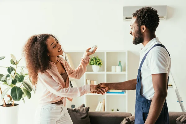 Happy african american woman holding air conditioner remote controller and shaking hands with african american handyman — Stock Photo