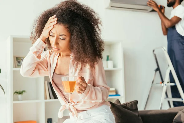 Femme afro-américaine épuisée avec tasse de thé tenant la main sur la tête, tandis que réparateur afro-américain fixant climatiseur — Photo de stock